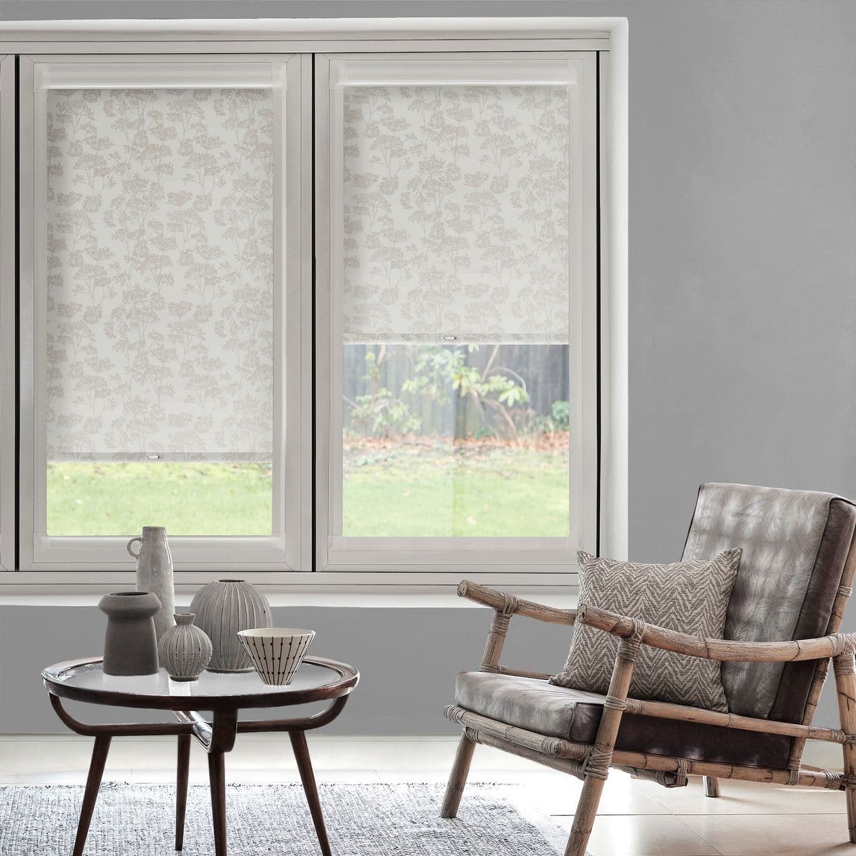 Floral-patterned roller blinds covering twin windows, partially lowered with translucent lower panels; filtering soft daylight into a grey-walled living room with rattan armchair, patterned cushion and round coffee table.