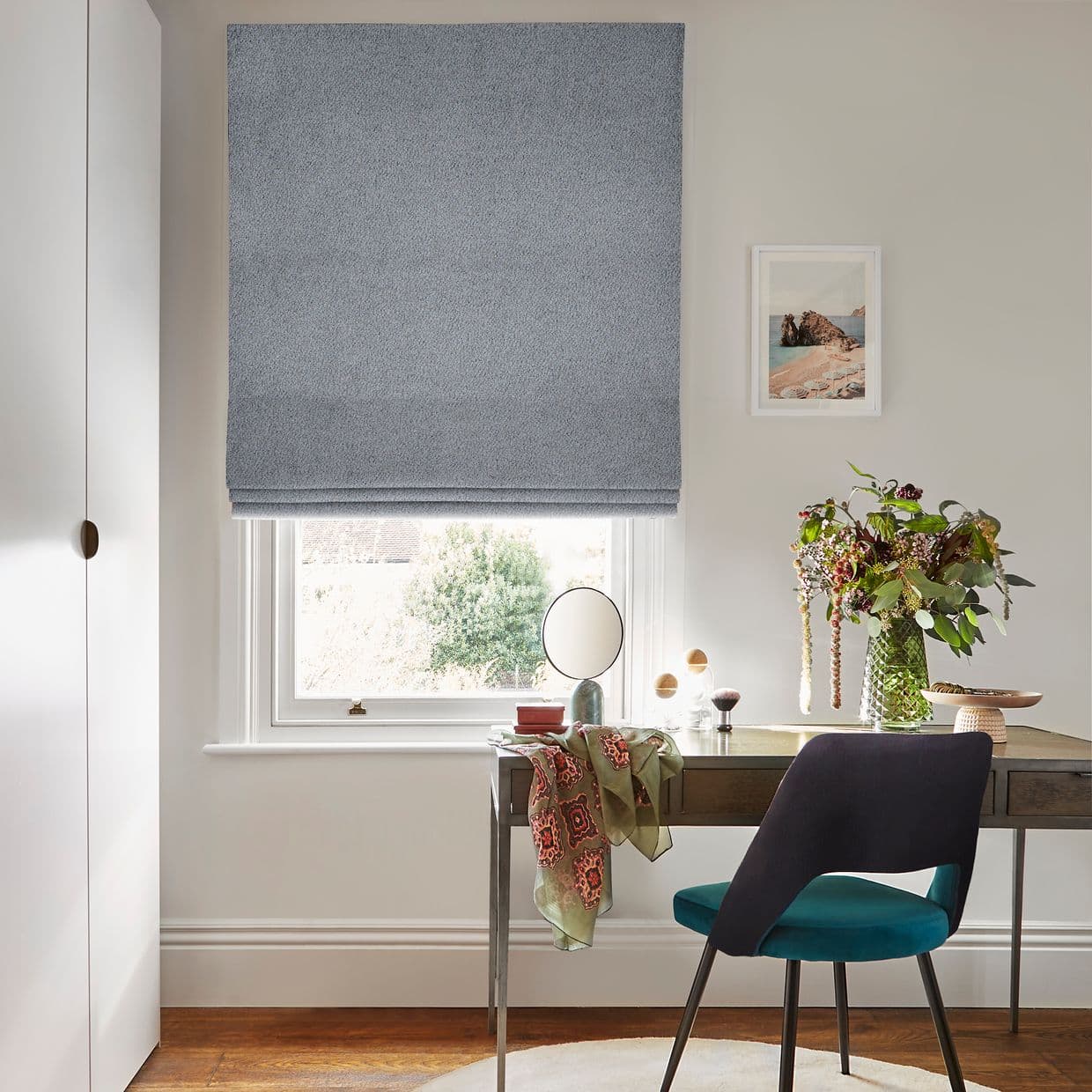 A textured light-blue Roman blind, lowered to mid-window with soft folds, gently filtering daylight; in a bright home office, desk, teal chair, vase of flowers and framed coastal print nearby.