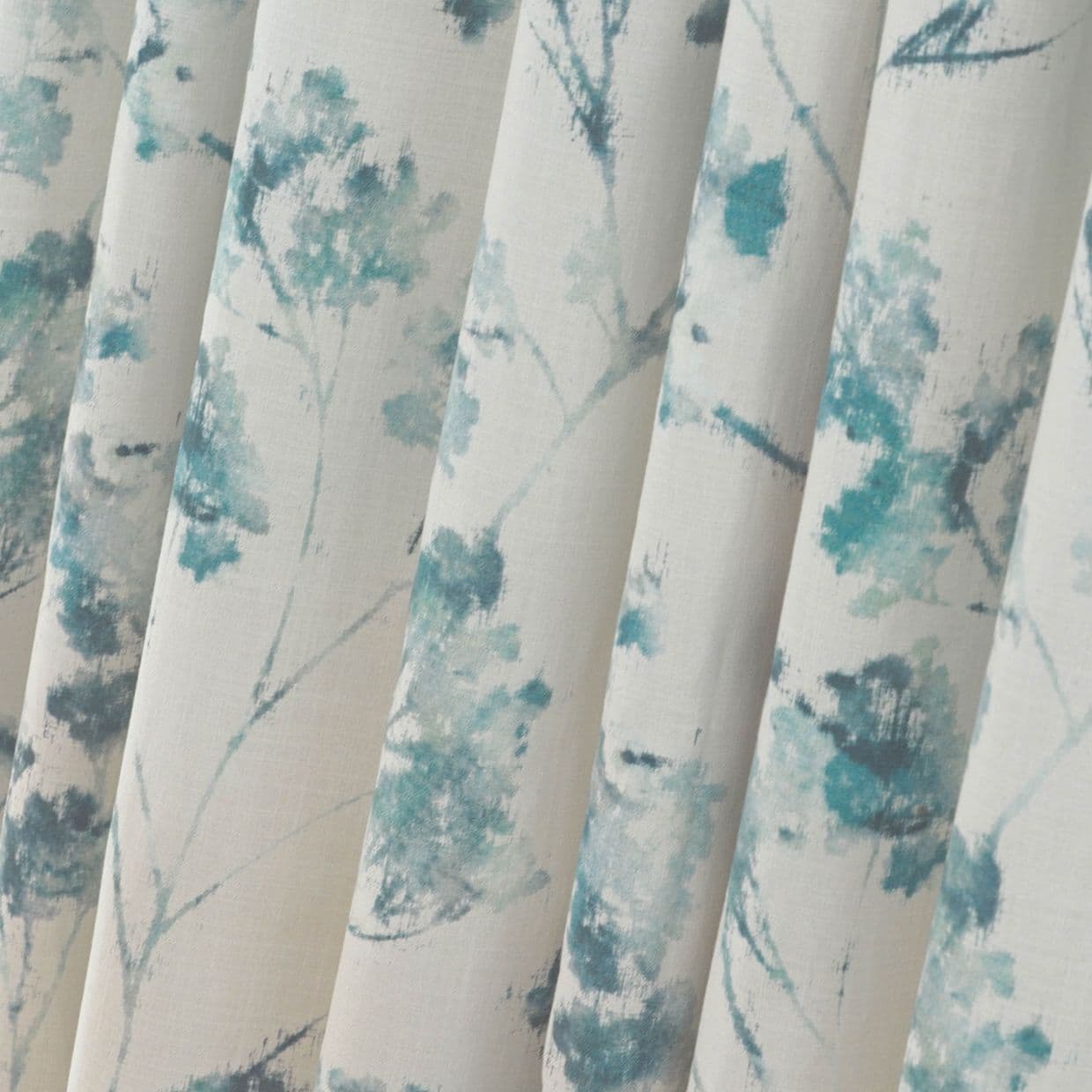 Fabric curtains with vertical pleats and teal watercolor floral print, hanging in soft folds; close-up interior view with neutral cream background and diffuse natural light.