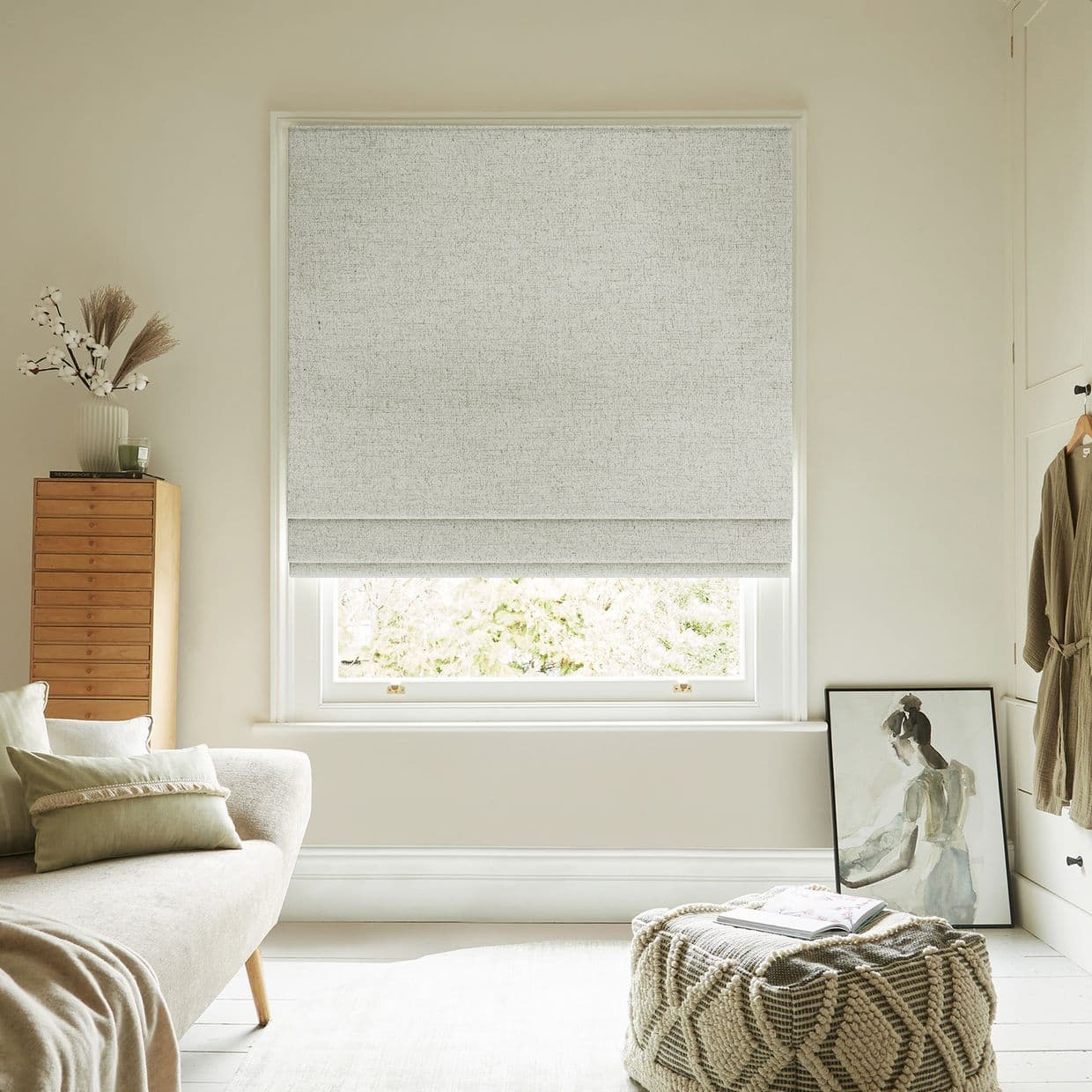 Light-gray textured Roman blind lowered with two soft folds, filtering diffuse daylight; in a neutral, cozy living room with sofa, wooden chest, knitted pouf and a leaning framed painting.