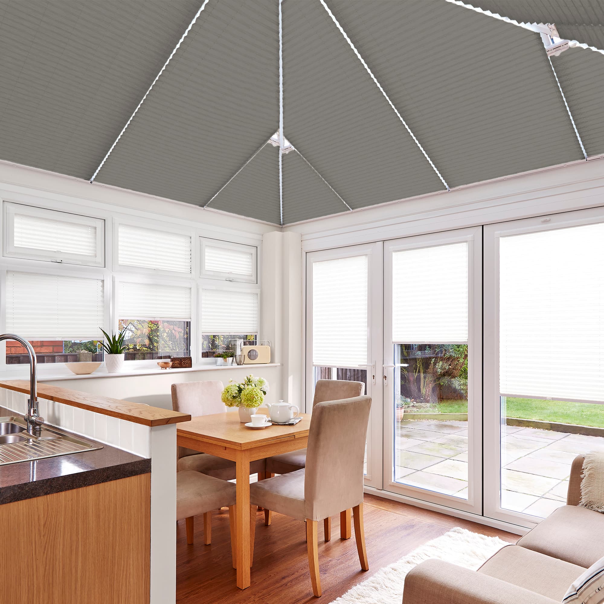 White pleated cellular blinds cover windows, French doors and pitched roof panels; partially drawn, filtering daylight into a bright dining sunroom with wooden table, beige chairs and garden view.