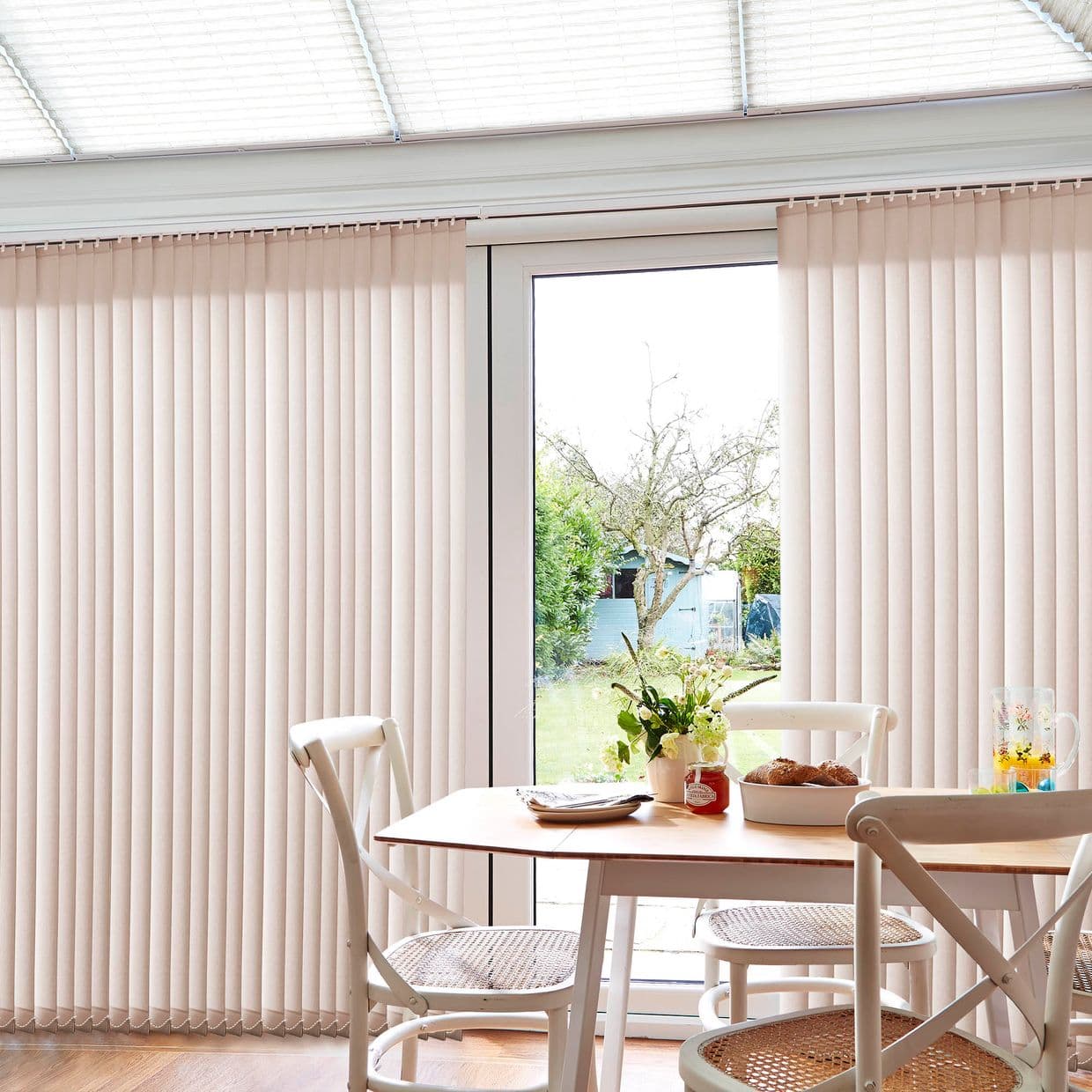 Beige vertical blinds covering a sliding glass door, parted centrally to reveal a garden; filtering bright daylight into a small dining area with wooden table, cane-seat chairs, flowers and pastries.