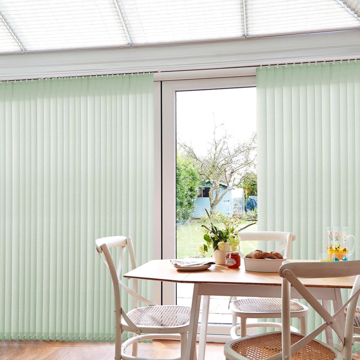 Pale-green vertical blinds hang from a top track, drawn to sides to expose a central glass door while filtering light; dining table and chairs sit in a bright, garden-facing room.