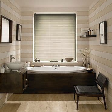 Horizontal venetian blind inside central window, closed and filtering soft daylight; in a neutral-striped bathroom with a recessed bathtub in dark wood surround, vessel sink, mirrors, shelf and a chair.