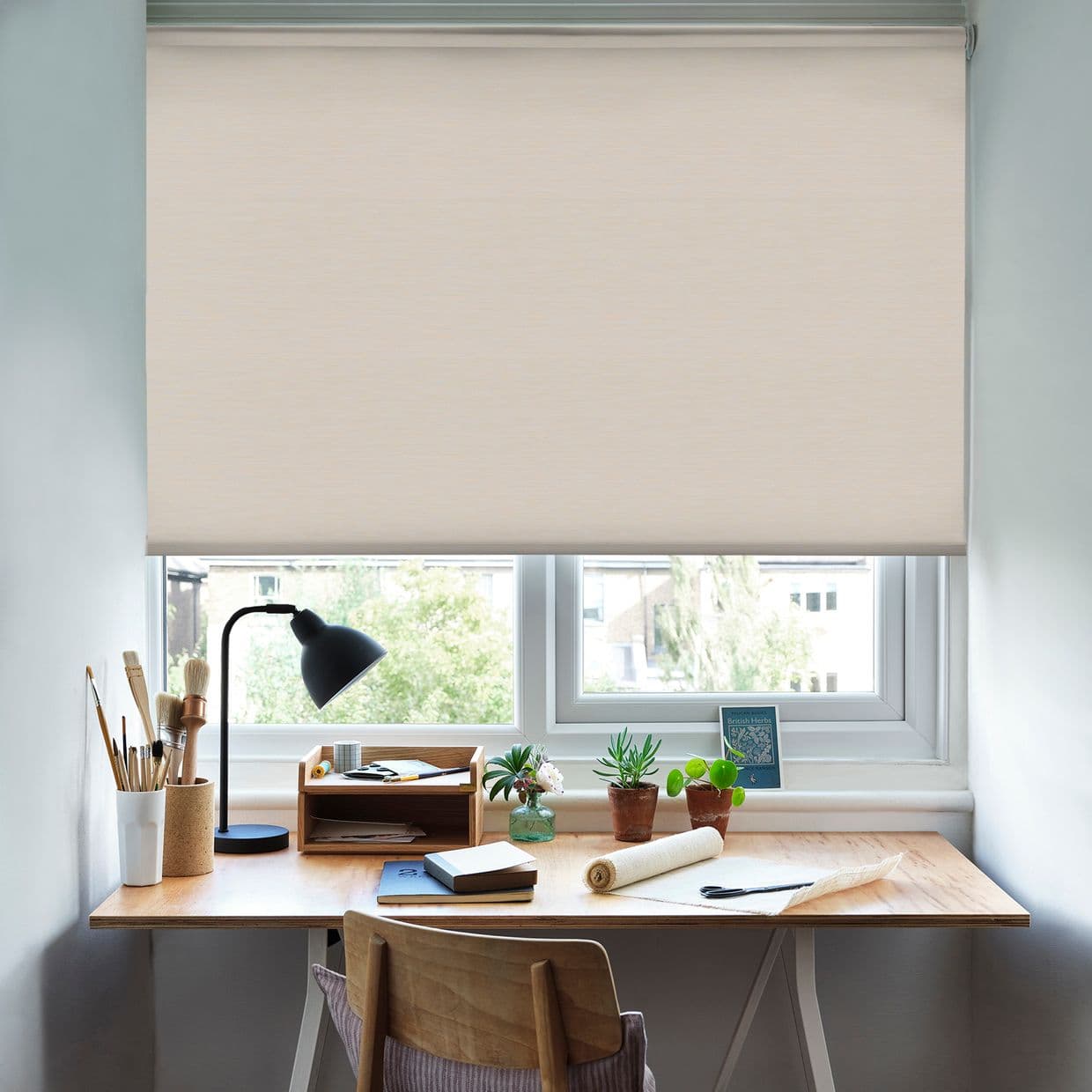 Beige fabric roller blind lowered from a top cassette, softly filtering daylight and blocking most of the window; home desk beneath with lamp, notebooks, brushes, potted succulents and wooden chair.