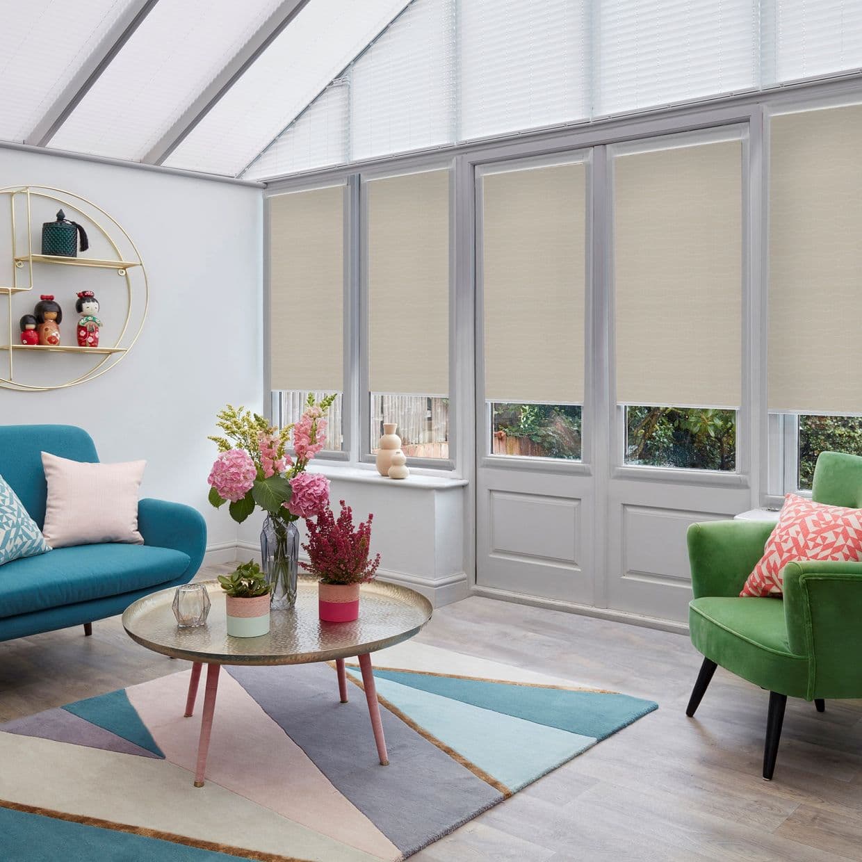 Beige roller blinds on tall conservatory windows/doors, partially lowered to obscure upper panes and filter daylight; bright sitting room with skylights, teal sofa, green armchair, and round coffee table.