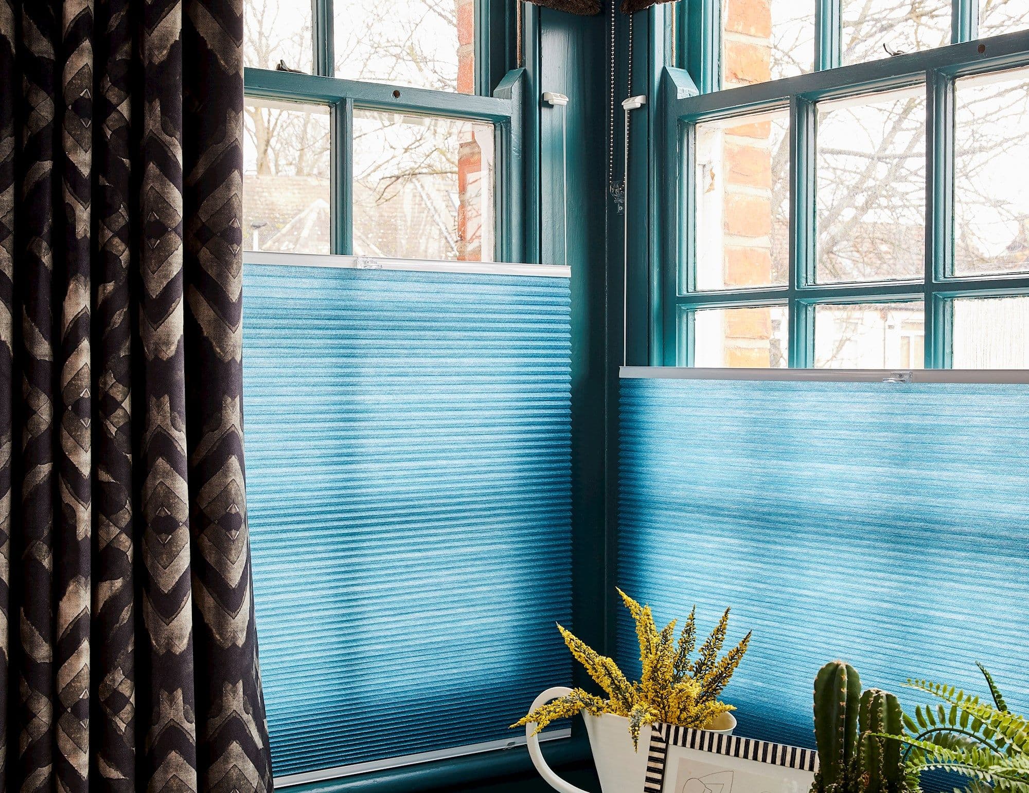 Dark geometric curtain hangs left while turquoise pleated shades cover lower panes, filtering daylight; teal window frames reveal bare trees and brickwork outside, with potted plants on the sill.
