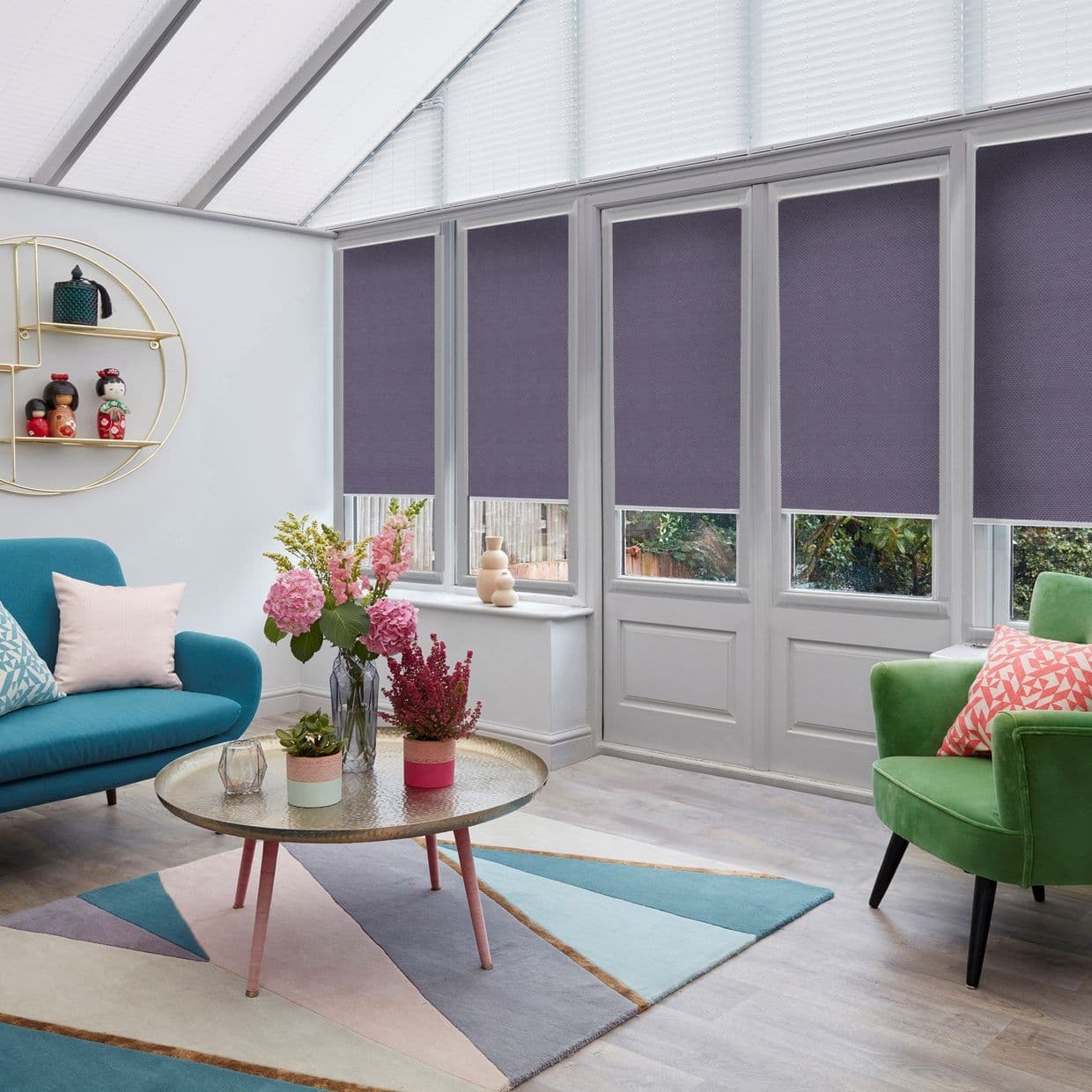 Plum roller blinds on tall windows and a door, lowered leaving narrow clear lower panes; filtering daylight in bright conservatory with pitched translucent roof, teal sofa, green armchair, pastel rug.