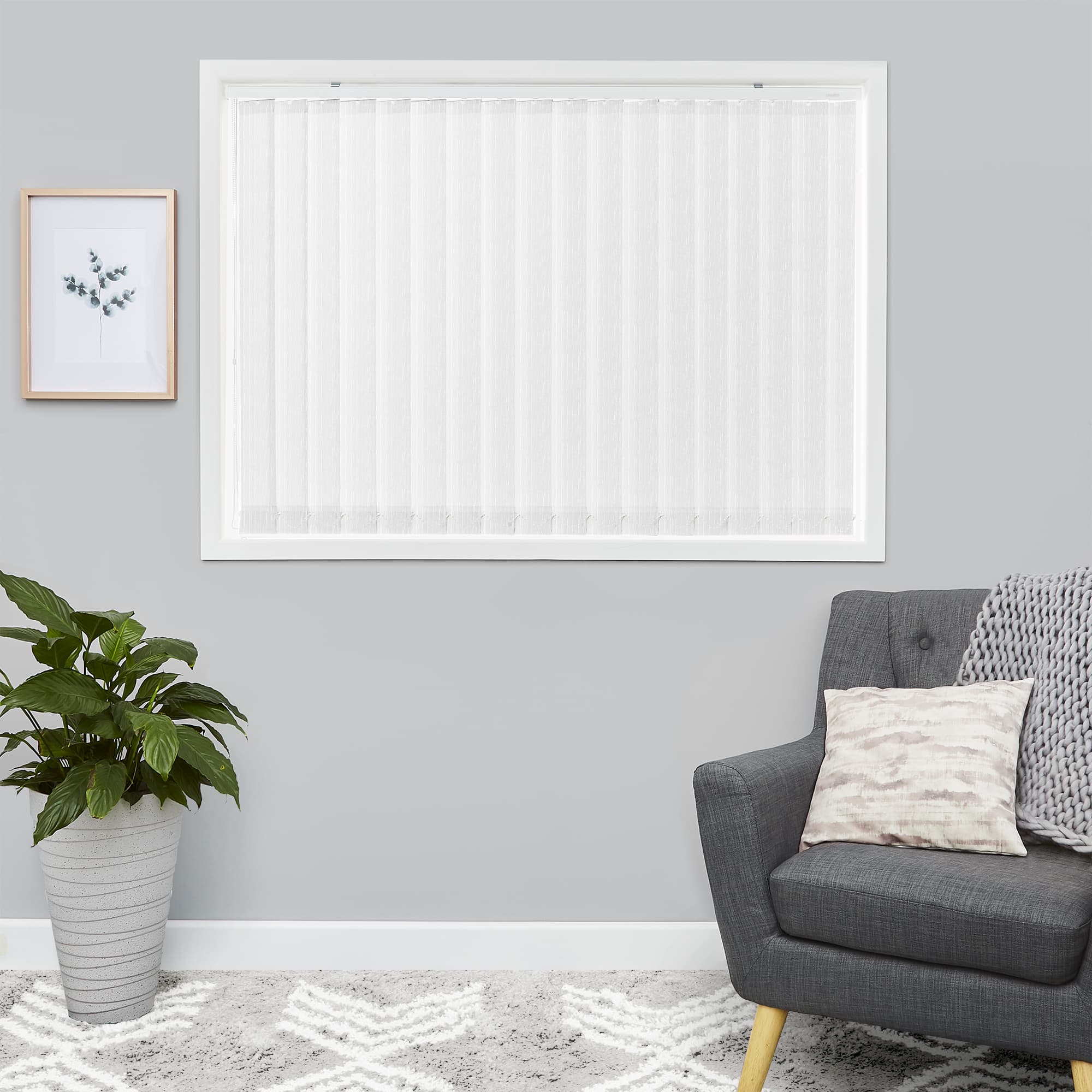 White vertical blinds covering the window, closed and softly diffusing daylight; set in a gray living room with a gray upholstered armchair, patterned rug, potted plant, and framed botanical print.