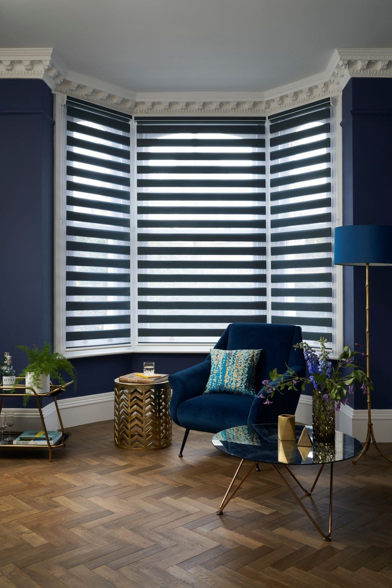Layered horizontal zebra blinds covering a three-panel bay window, alternating opaque and sheer bands, partially filtering daylight; set in a navy living-room corner with velvet armchair and parquet floor.