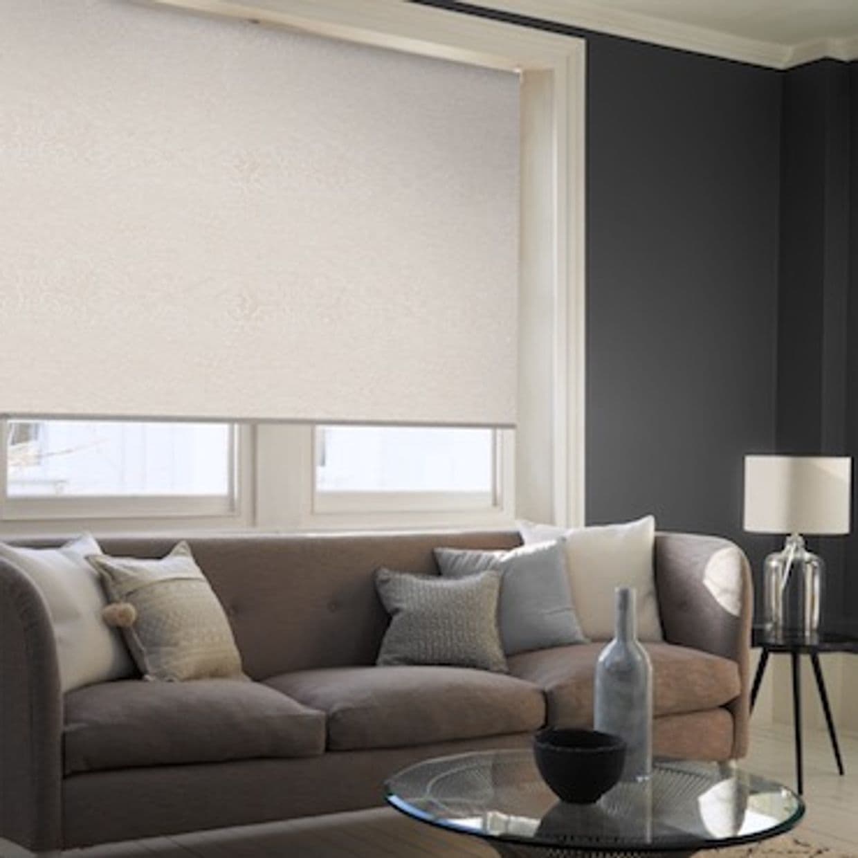 A light beige roller blind covers a double window, fully lowered and diffusing daylight; modern living room with dark grey walls, tufted sofa, several cushions, glass coffee table and side lamp.