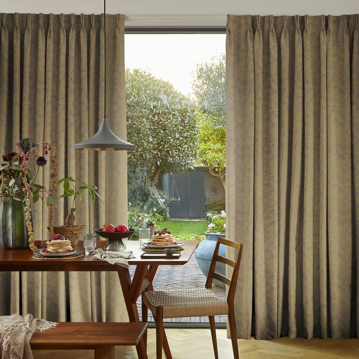 Heavy pleated patterned floor-to-ceiling curtains, partially drawn to sides, framing a central glass door; filtering soft daylight into a dining nook with wooden table set for breakfast and garden view.