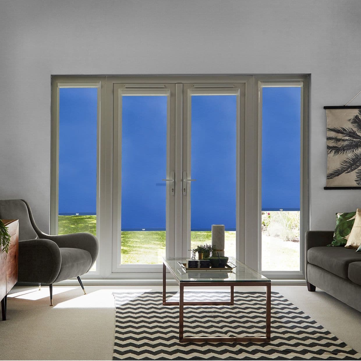 Blue roller blinds across double French doors and narrow sidelights, fully pulled down blocking view and filtering daylight; modern living room with grey armchairs, glass coffee table, chevron rug, lawn glimpsed below.