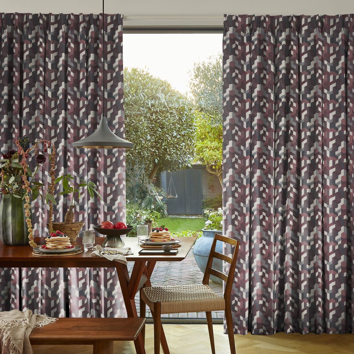 Patterned pleated curtains with geometric maroon and grey blocks, pulled aside, framing an open glass patio door and filtering daylight; dining table with breakfast setting and garden view.