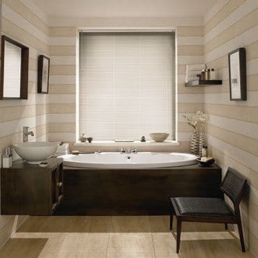 Horizontal Venetian blinds (pale, fully lowered) cover the central window, slats filtering soft daylight; they sit above a dark-wood built-in bathtub in a beige-striped, warmly lit bathroom.
