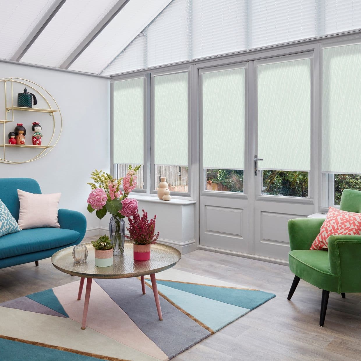 Pale mint, textured roller blinds lowered mid-height across tall conservatory doors and windows, diffusing daylight; cozy living room with teal sofa, green armchair, geometric rug, round coffee table with flowers.