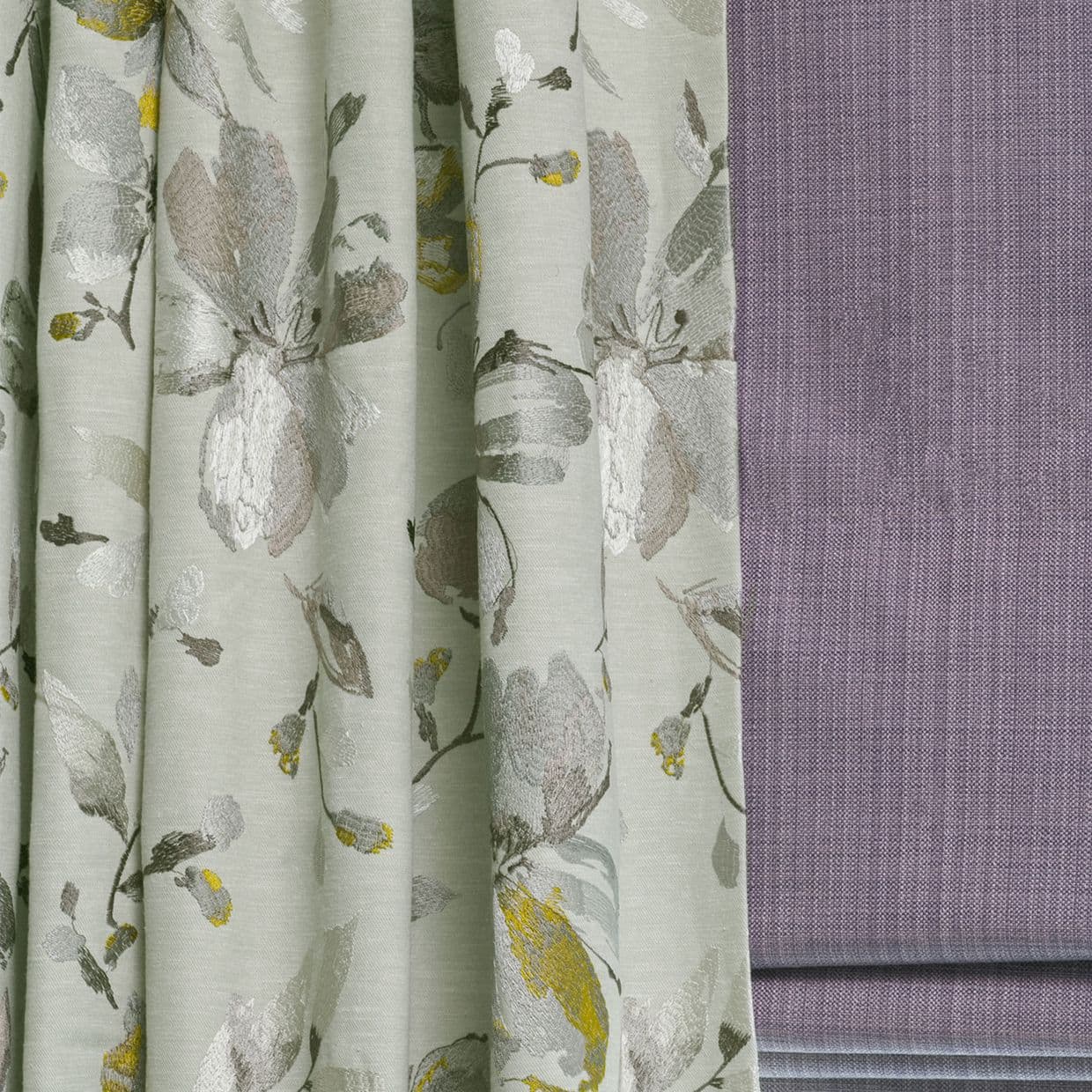 Floral jacquard curtains (left) with soft vertical pleats, hanging and slightly gathered; textured mauve Roman blind (right) folded and lowered; close-up of fabrics in a softly lit interior.