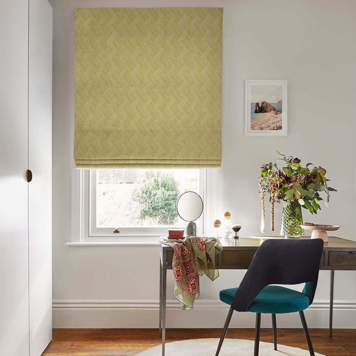 Yellow-green herringbone Roman blind, lowered mid-window and filtering soft daylight; small home desk setup with teal chair, round mirror, vase of flowers, pale walls and garden view.
