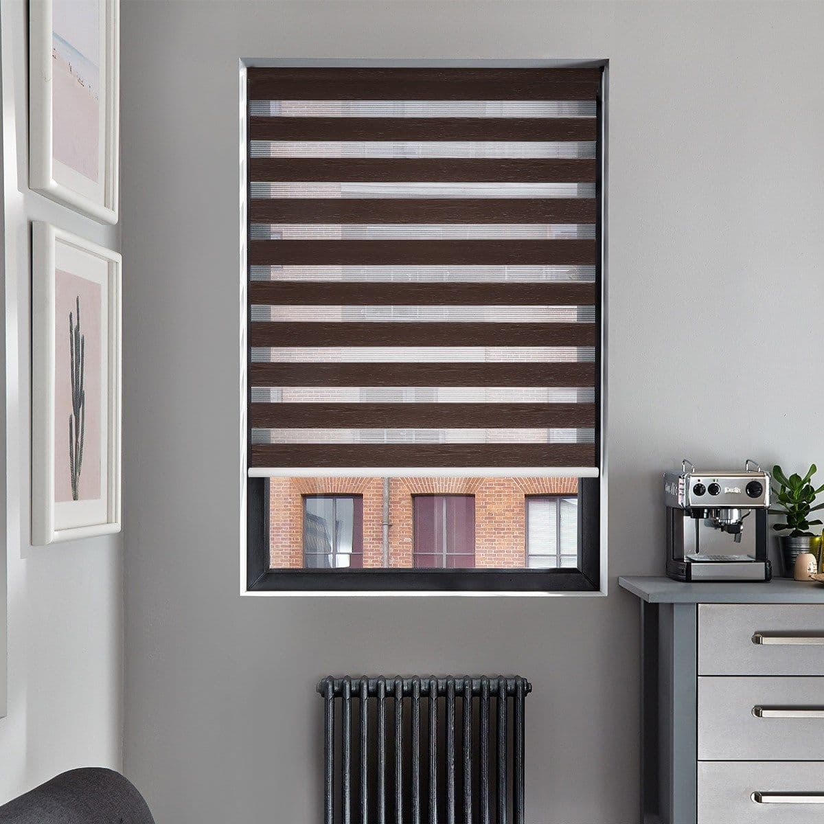 Brown zebra roller blind, lowered with alternating opaque and sheer horizontal stripes, bottom raised; filtering daylight and revealing a brick-building view in a gray kitchen with radiator and coffee machine.