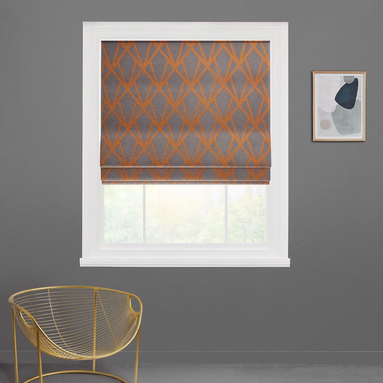 Roman shade with gray fabric and orange geometric lattice, lowered over a white window with its bottom raised, filtering daylight; gray wall, gold wire chair left, abstract print right.