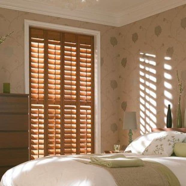 Wooden plantation shutters covering a tall windowâ€”four full-height panels with adjustable slatsâ€”partially closed, casting striped sunlight across a softly lit bedroom with bed, nightstand, patterned wallpaper and dresser.