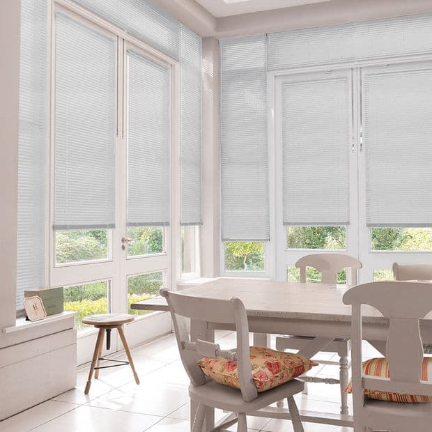 White pleated blinds covering tall French windows and patio doors, lowered to mid-height and filtering daylight; bright dining/sunroom with wooden table, chairs with floral cushions, tiled floor, garden view.