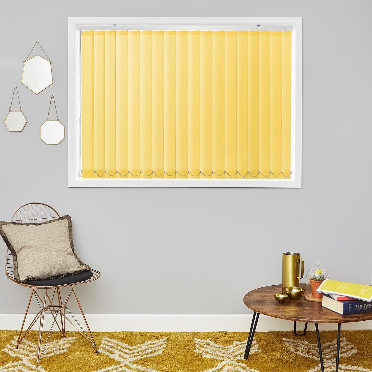 Vertical yellow blinds covering a white-framed window, closed and diffusing light; mounted on a grey wall above a mustard rug, beside a wire chair and a wooden coffee table.
