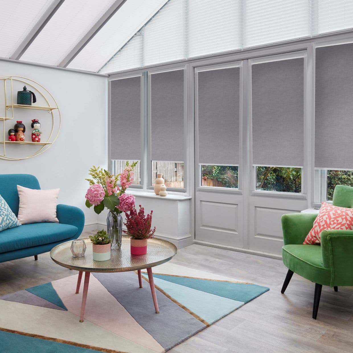 Grey roller blinds, lowered to cover most of four tall panes, leaving narrow lower openings exposed, partially blocking light; bright conservatory living room with colorful sofas, rug, and flowers.