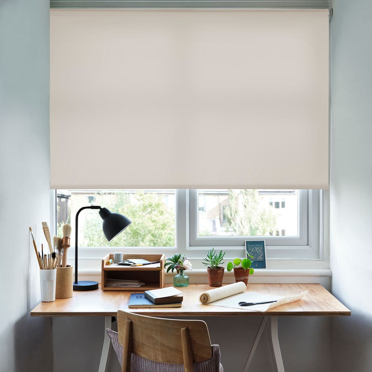 A cream roller blind, lowered almost fully and softly diffusing daylight, covers a double window above a wooden desk with lamp, art brushes, potted plants, notebooks and chair, suburban view.