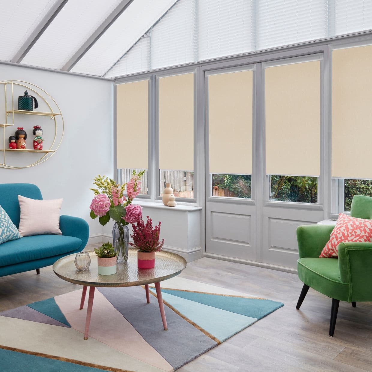 Beige roller blinds lowered over tall doors and windows, covering upper panes and diffusing daylight; in a bright conservatory living room with teal sofa, green armchair, coffee table and flowers.
