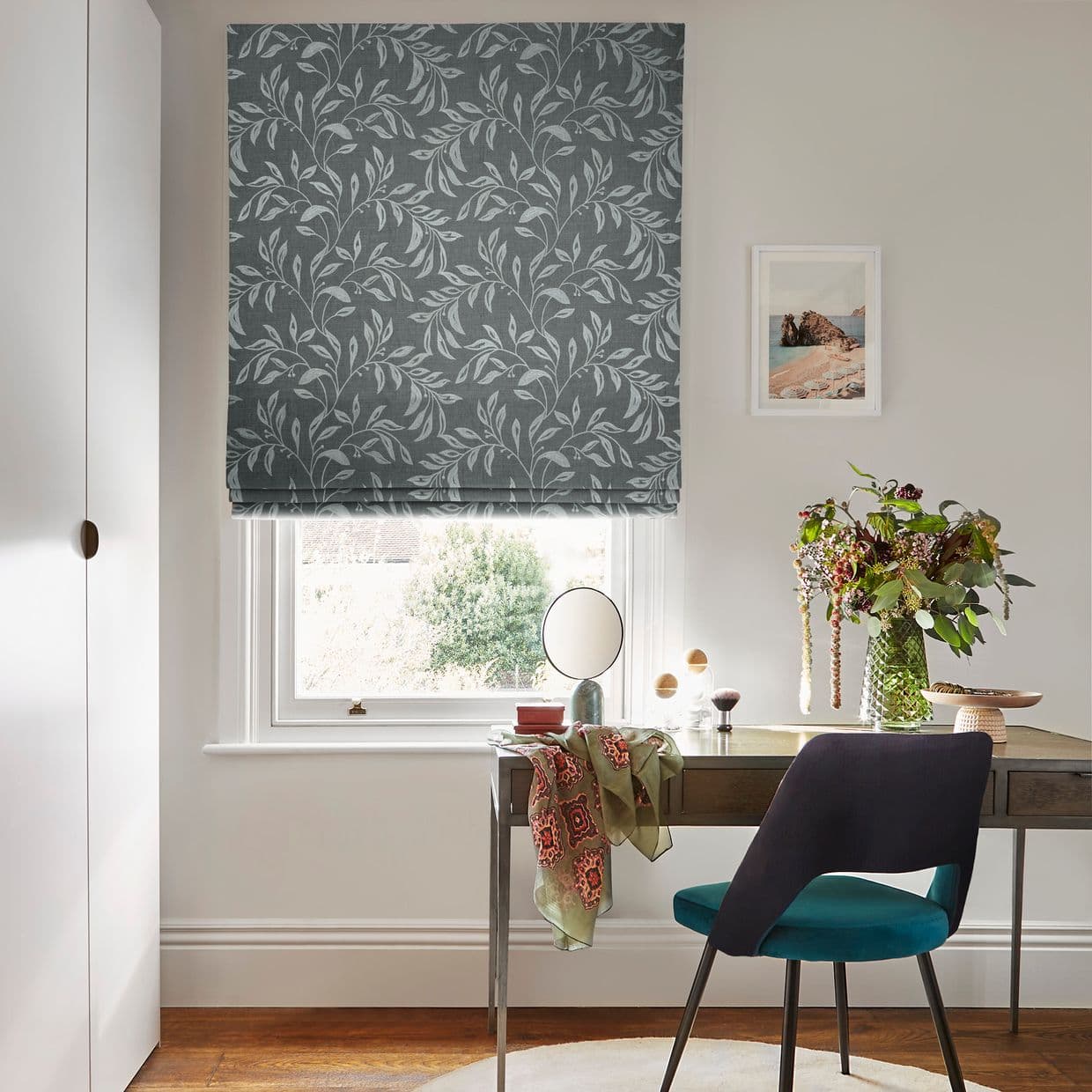 Grey floral Roman blind, lowered in soft folds, partly filtering daylight with a narrow gap at the bottom; positioned over a window above a desk in a bright home office.