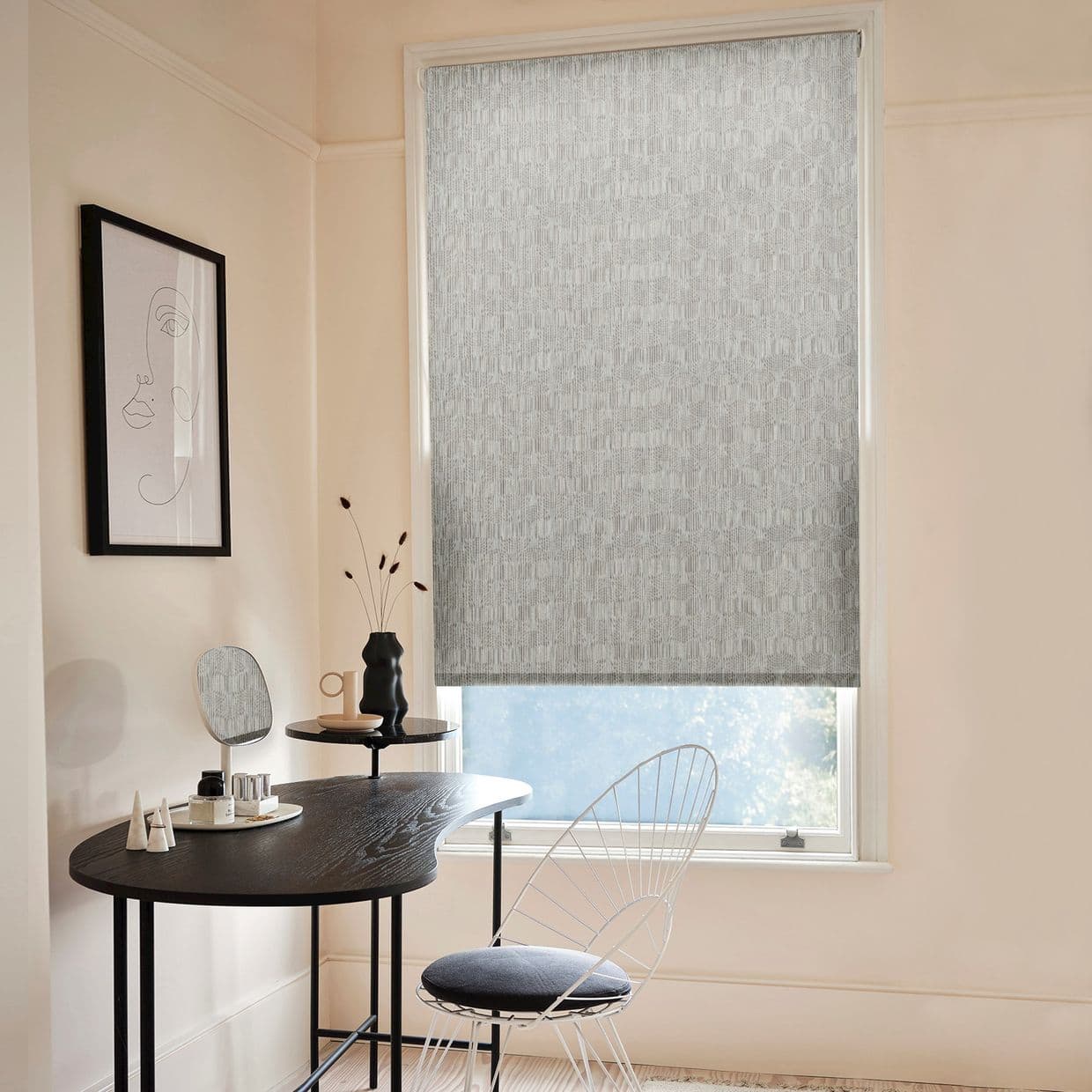 A grey patterned roller blind, lowered over a tall window, filtering daylight â€” in a pale minimalist dressing nook with a black desk, wire chair and framed line-art.