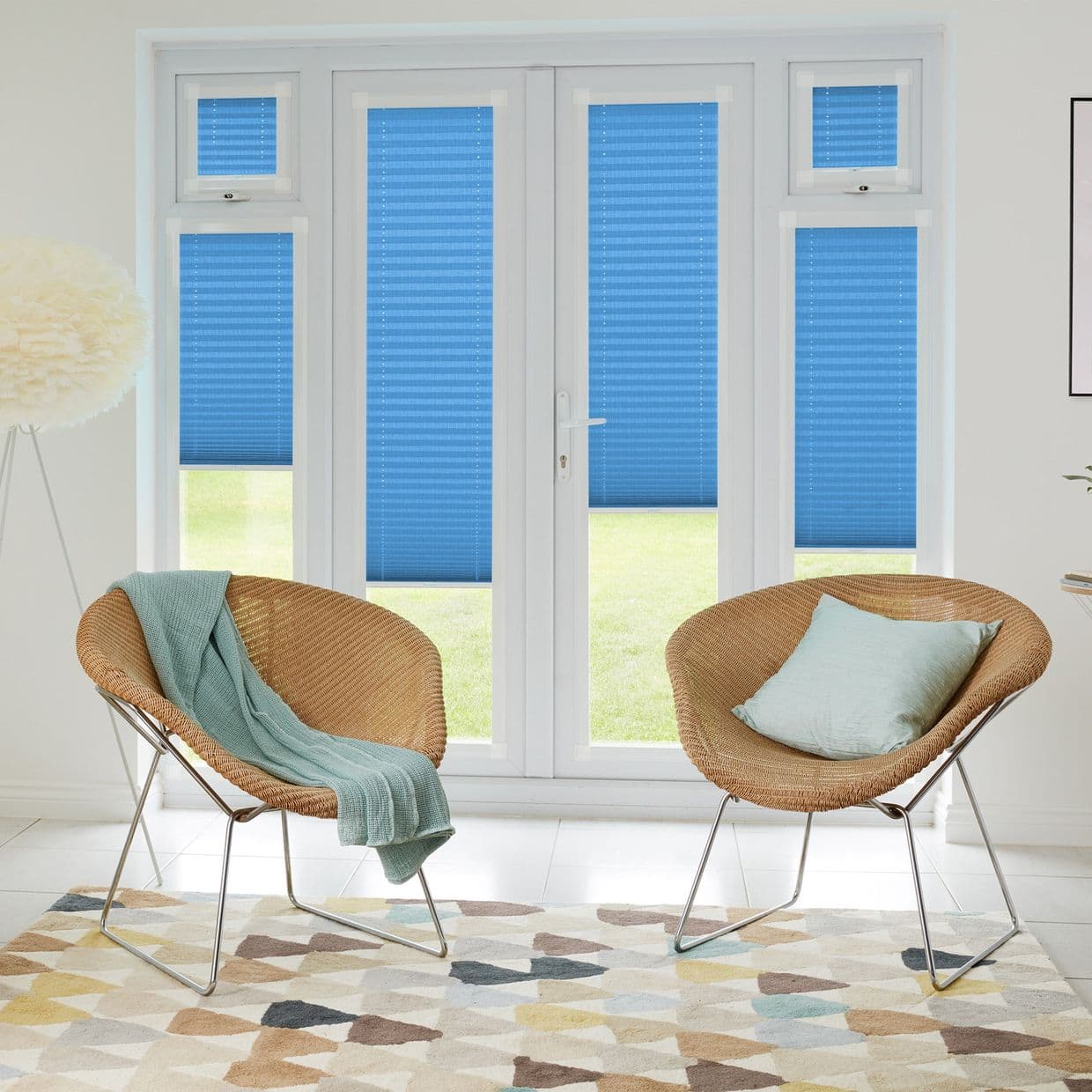 Blue pleated blinds cover French doors and narrow sidelightsâ€”mostly lowered, two panels partially raisedâ€”filtering daylight into a bright sitting area with two wicker chairs, a throw, cushion, and patterned rug.