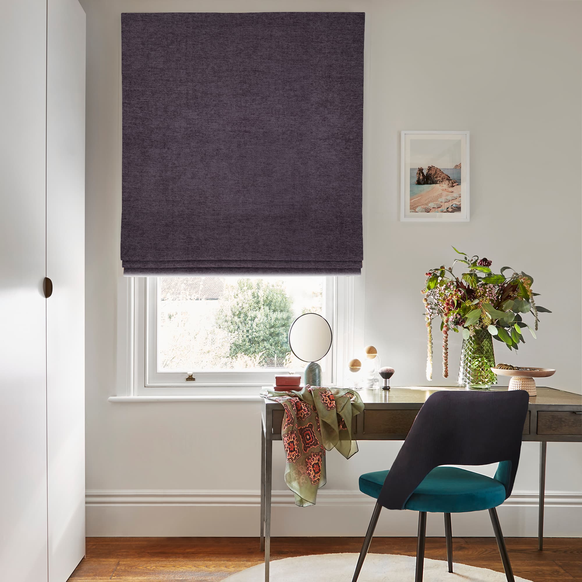 Roman fabric blind, deep plum, lowered with soft folds near the bottom; filtering daylight. Context: small home study â€” wooden desk with mirror, vase of foliage, teal-upholstered chair and wall art.