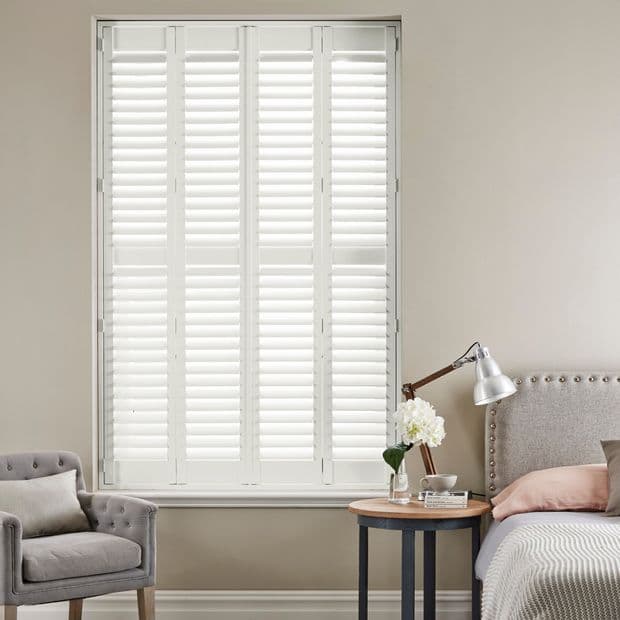 White full-height plantation shutters (four panels with adjustable louvers) covering the window; closed, louvers angled to diffuse bright daylight; neutral, softly lit bedroom with upholstered bed, side table, lamp, armchair.