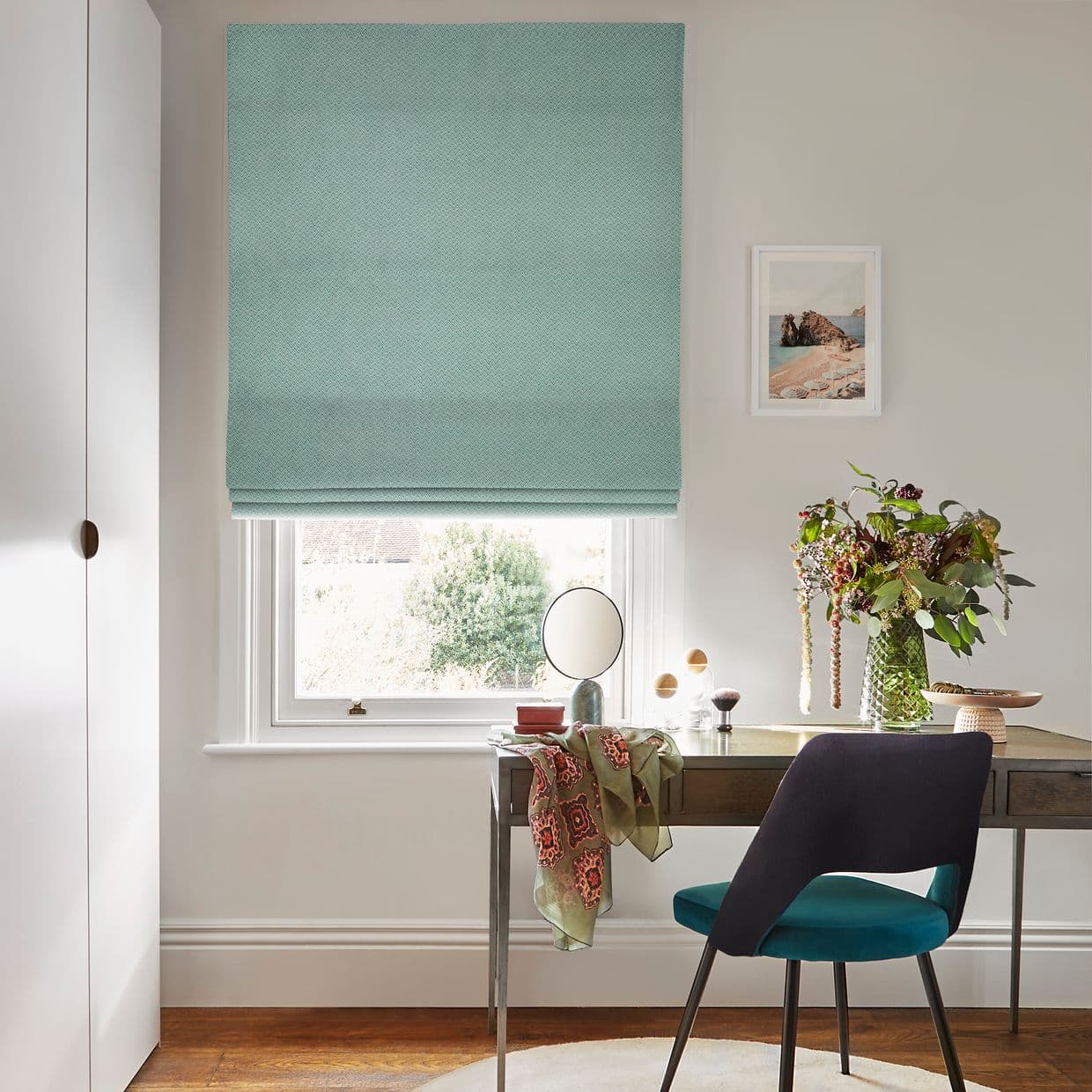 Teal patterned Roman blind, partially lowered and filtering daylight over a sash window above a desk in a bright home office with a teal chair, vase of flowers and mirror.