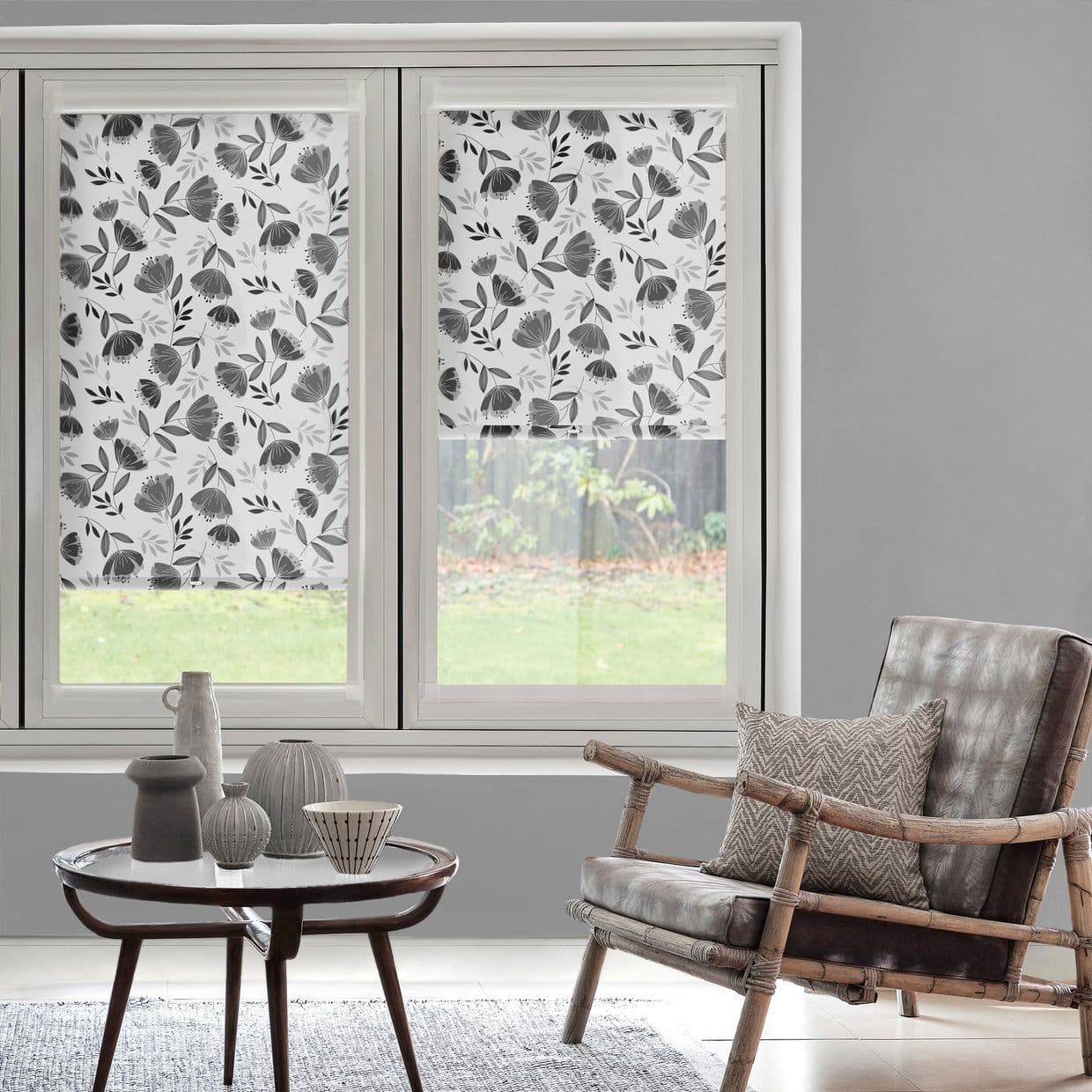 Floral roller blinds (white with gray botanical print) partially lowered on two adjacent windows, filtering daylight and obscuring view; modern living room with rattan armchair, cushion, coffee table and vases.