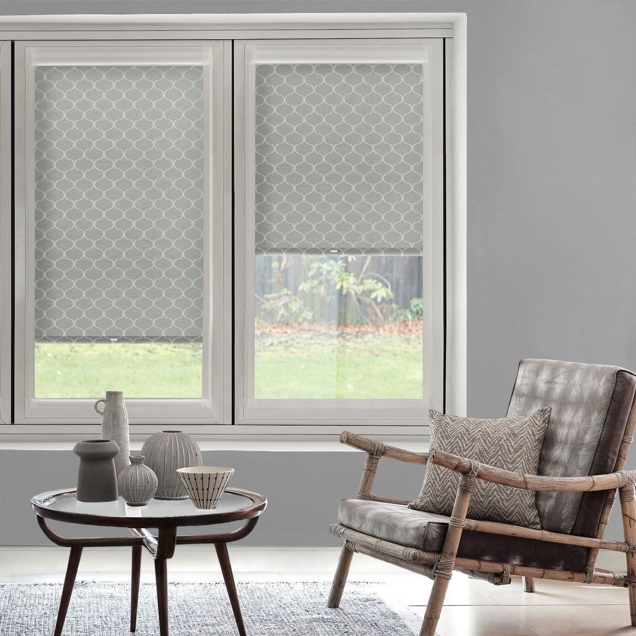 Patterned roller blinds on twin windowsâ€”one lowered, one halfâ€‘raisedâ€”filtering light through grey geometric fabric; in a neutral living room with a wooden coffee table and rattan armchair in soft daylight.