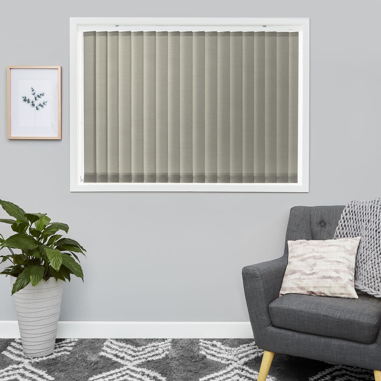 Fabric vertical blinds cover a centered rectangular windowâ€”beige-gray slats, fully closed and filtering soft daylight; gray-walled living room with a potted plant, framed botanical print, gray armchair and patterned rug.