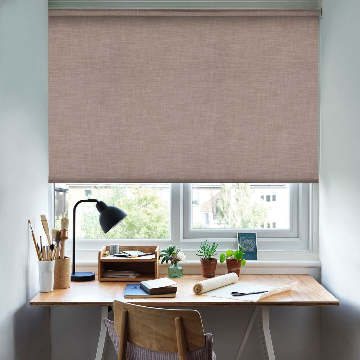 A textured, pale pink fabric roller blind lowered over a double window, filtering soft daylight; beneath it a wooden home-study desk with lamp, brushes, notebooks, small potted plants and chair.