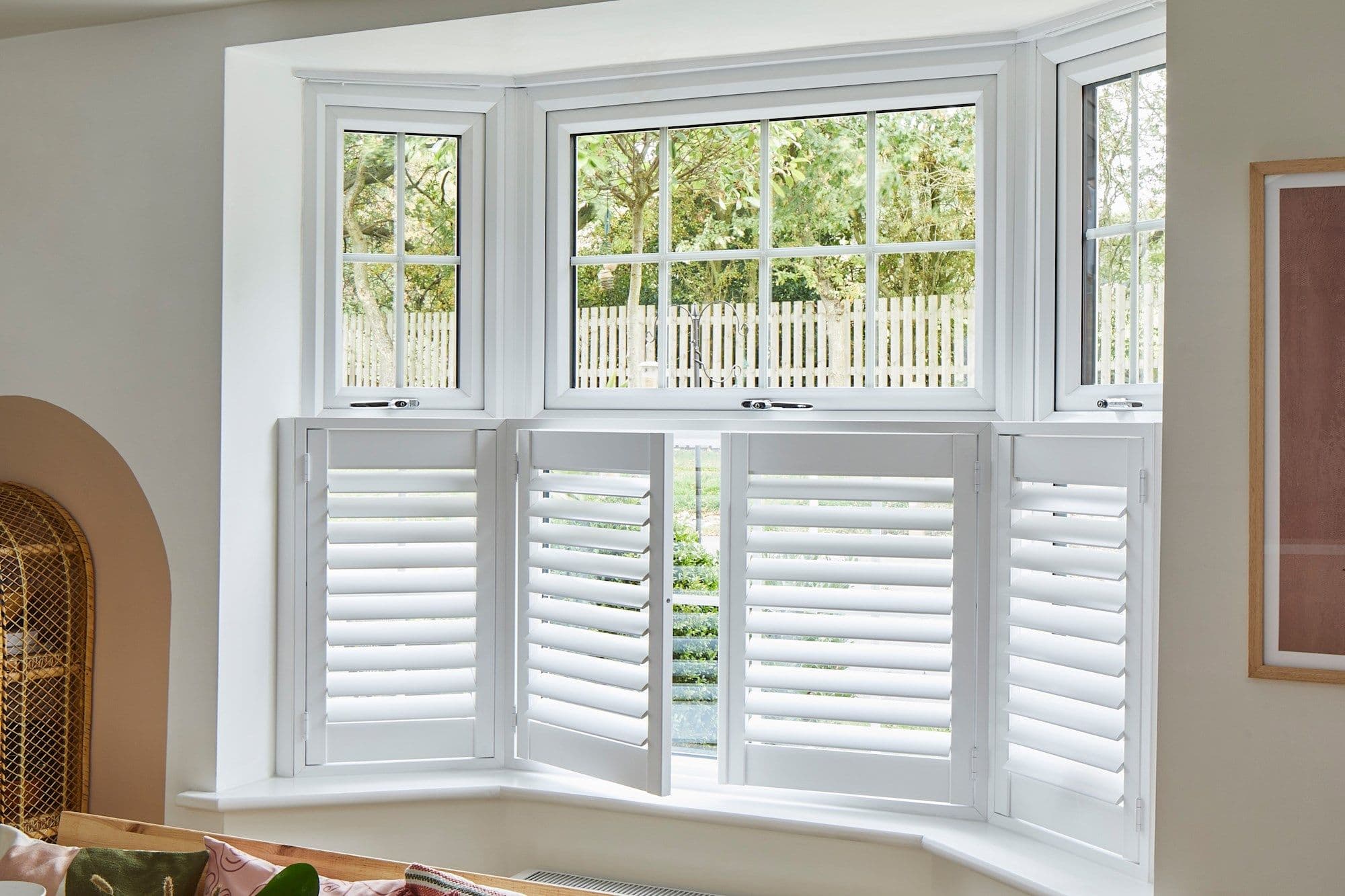 White plantation shutters (half-height, hinged panels) across a three-panel bay window, one panel ajar; slats tilted to filter daylight into a bright living room overlooking a fenced, tree-lined yard.