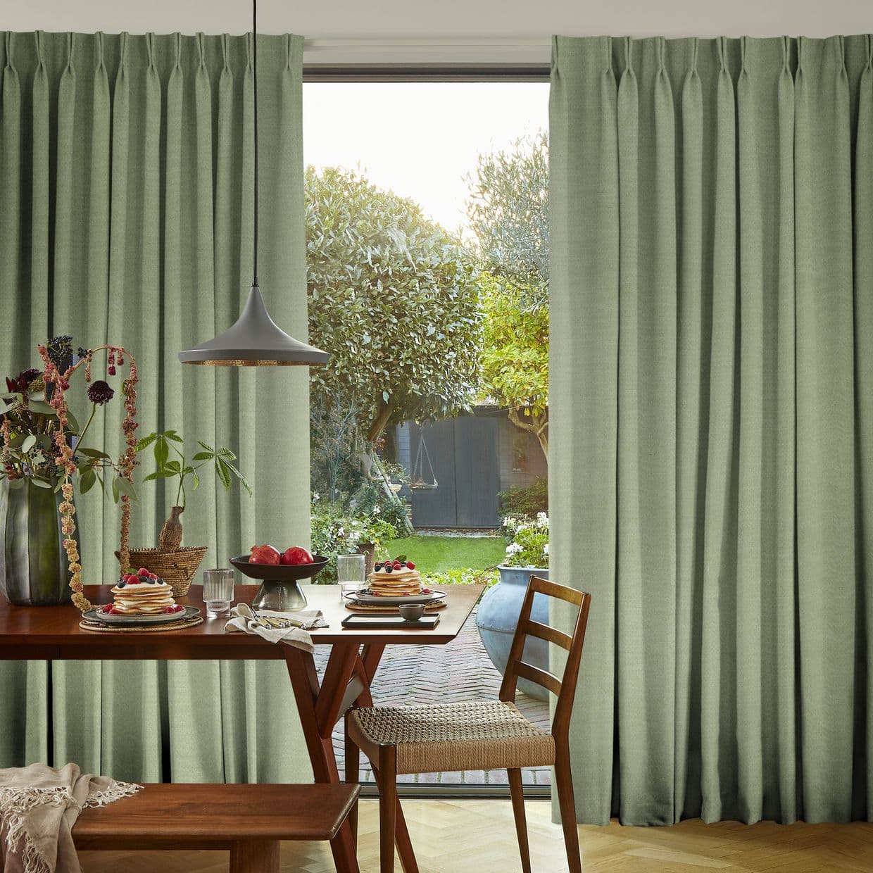 Floor-to-ceiling pleated sage-green curtains, partially parted, framing and softly filtering daylight onto a breakfast table set in a sunlit dining area opening onto a leafy garden.