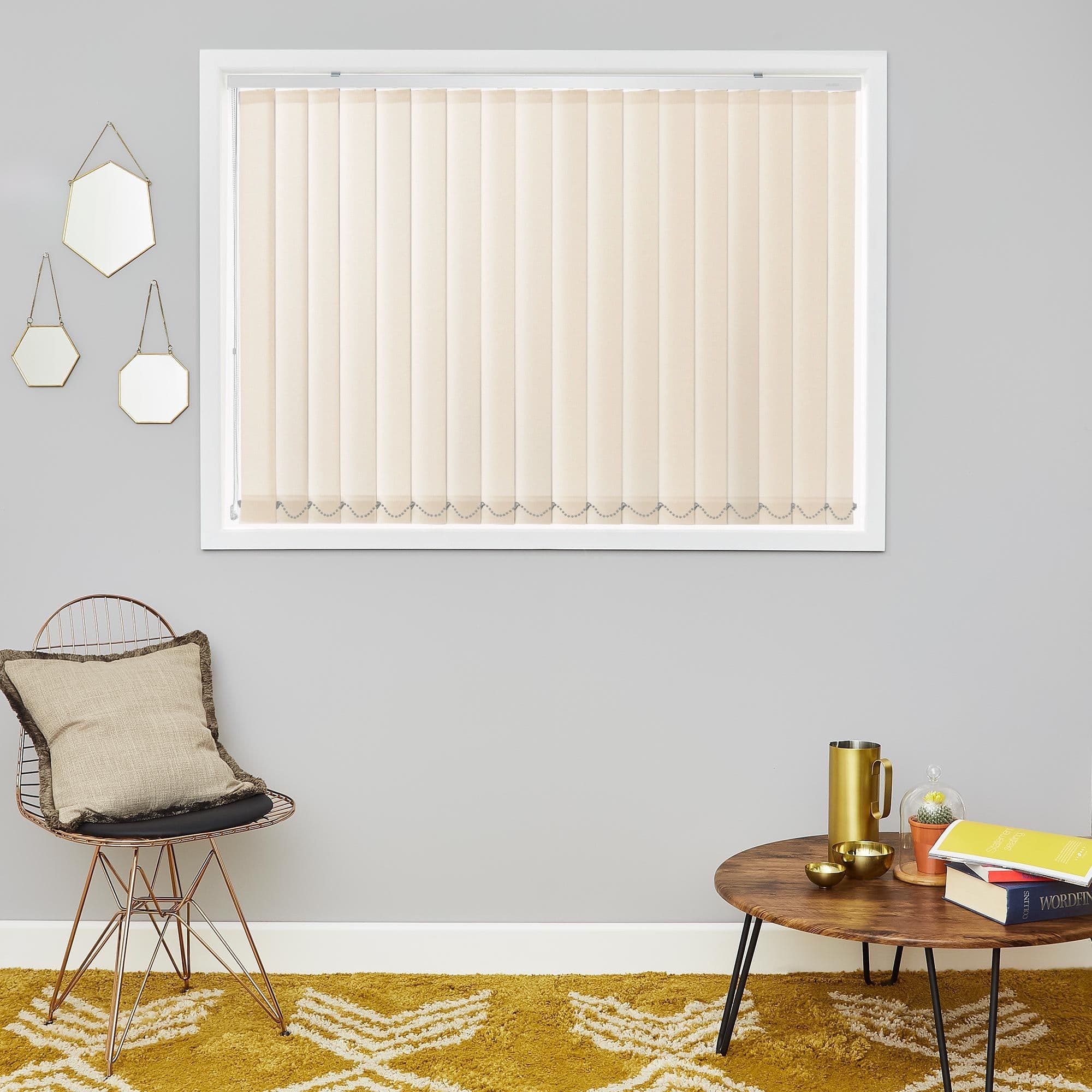 Beige vertical blinds covering a central rectangular window, closed with bead-chain weights along bottom, filtering soft daylight; minimal living room with gray walls, wire chair, wooden coffee table, mustard rug.