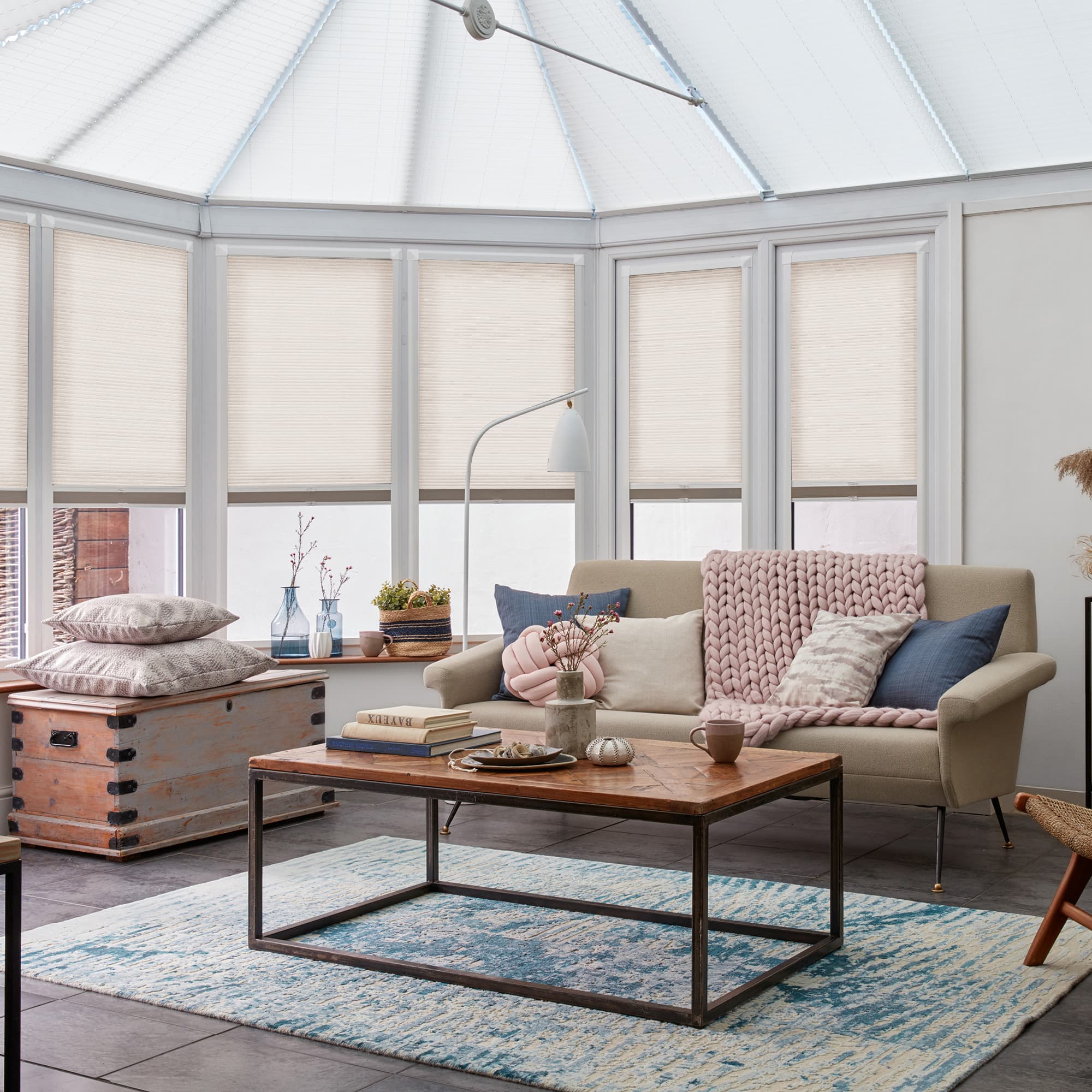 Off-white cellular (honeycomb) blinds lowered halfway on bay windows, gently filtering daylight; beige sofa with chunky pink throw faces wooden coffee table on blue rug in bright living area.