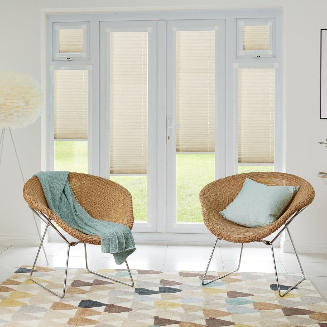 Beige pleated blinds on French doors and flanking windows, lowered to cover upper sections leaving clear lower panels, filtering daylight in living room with two wicker chairs and lawn view.