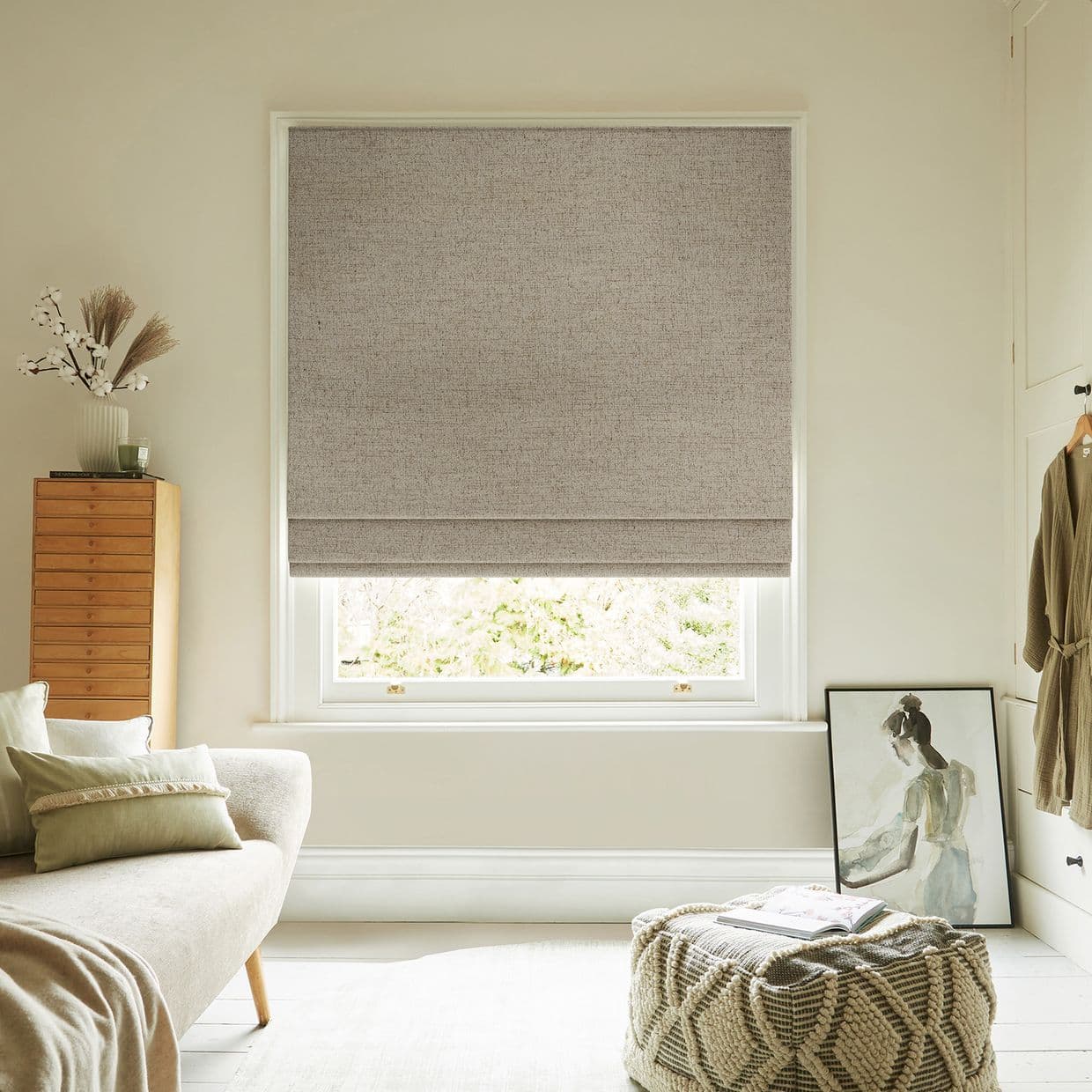 A full-width beige textured Roman blind lowered over the window, blocking view and softly filtering daylight; set in a neutral-toned living room with sofa, pouf, tall chest and leaning artwork.
