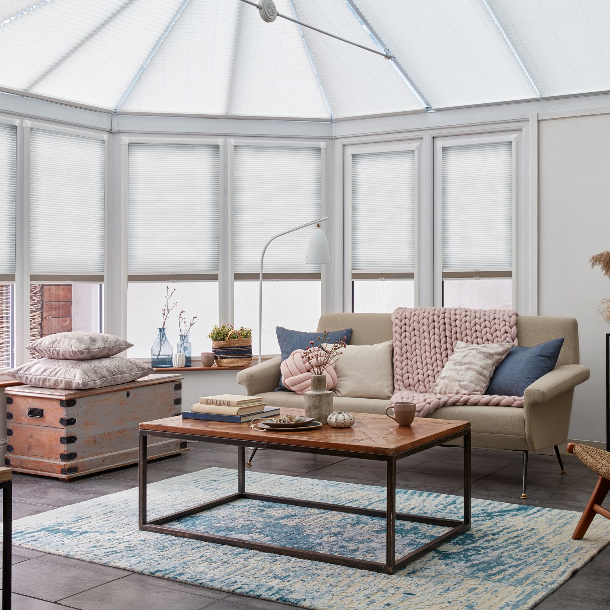 White pleated cellular blinds covering bay windows and glazed skylight, lowered to diffuse daylight; conservatory seating area with beige sofa, chunky pink throw, wooden coffee table and blue-pattern rug beneath.