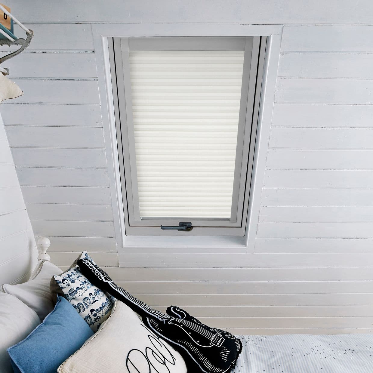 White pleated skylight blind, fully lowered with horizontal folds, filtering soft daylight; set in a white-painted attic with slatted ceiling above a bed with blue, patterned, and guitar-shaped pillows.