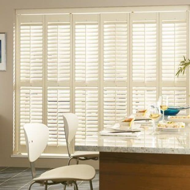 White louvered plantation shutters covering a full-height window; lower and upper panels closed with slats angled, filtering bright daylight into a modern kitchen/dining area with countertop and chairs.