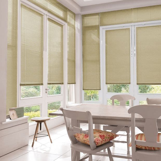 Beige pleated blinds lowered over tall white-framed French doors and windows, filtering soft daylight; a sunlit dining nook with pale wooden table, chairs and floral cushions, garden glimpses below.